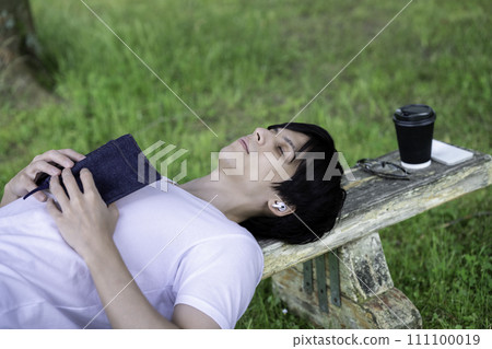 A foreign man lying on a park bench in fresh greenery, listening to music and reading a book. 111100019