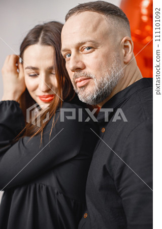 Romantic couple posing for a photo in anniversary. Studio full length shot of man and his wife near heart-shaped red ballon. Brunette woman and bearded man wearing black clothes. 111101092