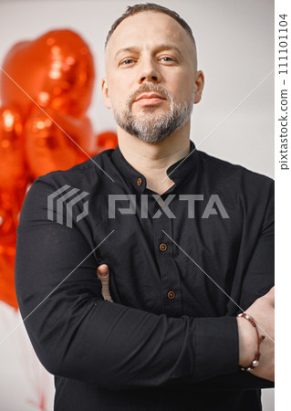 Man posing for a photo while standing in front. Studio full length shot man near bunch of heart-shaped red ballons. Brunette woman and bearded man wearing black clothes. 111101104