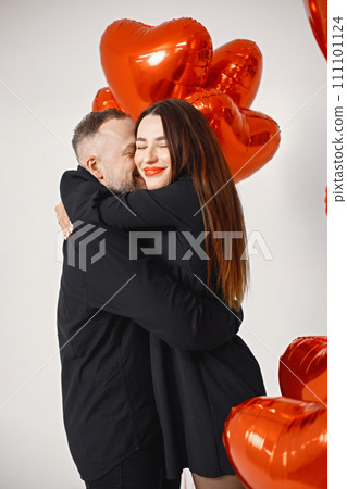 Romantic couple posing for a photo in anniversary. Studio full length shot of man embracing his wife and holding bunch of heart-shaped red ballons. Brunette woman and bearded man wearing black clothes 111101124