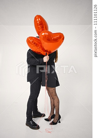 Romantic couple posing for a photo in anniversary. Studio full length shot of man kissing his wife and holding bunch of heart-shaped red ballons. Brunette woman and bearded man wearing black clothes. Romantic couple posing for a photo in anniversary. Studio full length shot of man kissing his wife and holding bunch of heart-shaped red ballons. Brunette woman and bearded man wearing black clothes. 111101126