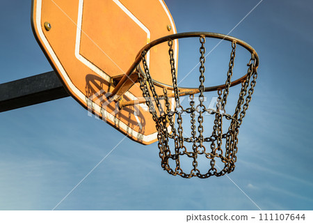 Side view of Basketball backboard with the hoop metal ring and steel chain net against blue sky background seen from below. Side view of Basketball backboard with the hoop metal ring and steel chain net against blue sky background seen from below. 111107644