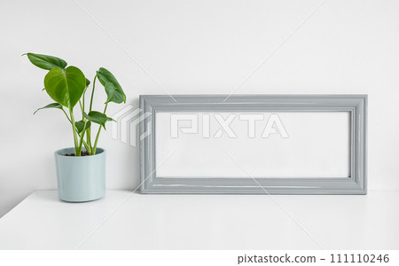 Gray mock up photo frame and Monstera deliciosa or Swiss Cheese Plant in a flower pot on a white table background Gray mock up photo frame and Monstera deliciosa or Swiss Cheese Plant in a flower pot on a white table background 111110246