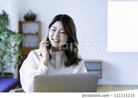 A young woman talking on a smartphone while using a laptop in the living room A young woman talking on a smartphone while using a laptop in the living room 111111215