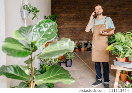 Floristry business and people concept. Male worker in uniform carrying flower in pot for customer and talking on mobile phone while working in flower shop. 111113434
