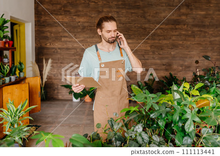 Caucasian male greenhouse worker in apron talks with clients on mobile phone during work in plant store or flower shop. 111113441