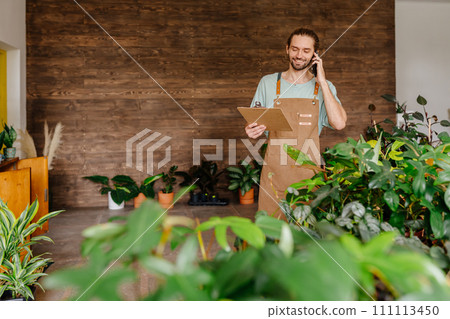 Smiling seller talking on smartphone and writing notes on clipboard in flower shop. Floristry business and people concept. 111113450