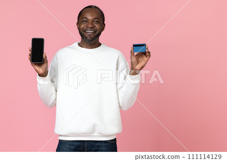 Cheerful black man wearing white sweatshirt showing phone with blank screen and credit card Cheerful black man wearing white sweatshirt showing phone with blank screen and credit card 111114129