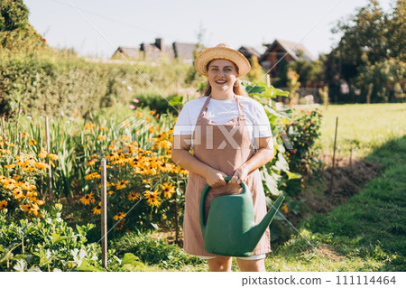 Beautiful happy young woman in a hat posing with green watering can in summer garden. Organic farm. Agriculture and farming concept. High quality photo 111114464