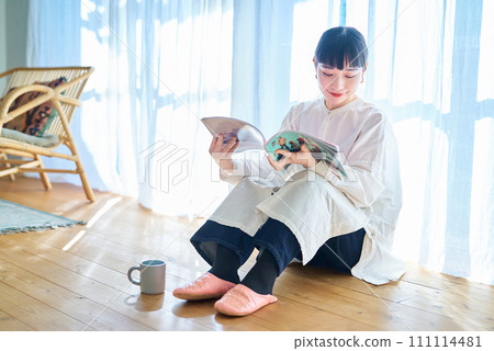 Young woman reading a magazine by the window in her room 111114481