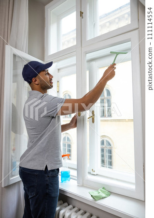 Young man in cap and T-shirt washing window at home with wiper squeegee. 111114554