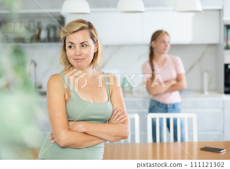 Upset woman standing in home kitchen after quarrel with teenage daughter 111121302