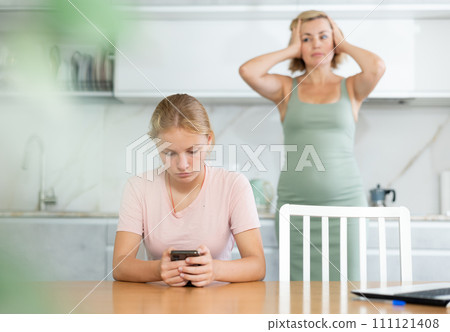 Teen girl fixated on phone while dissatisfied mother standing in background 111121408