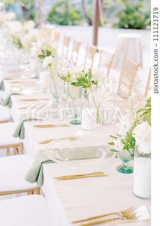Long set festive table with bouquets of flowers, transparent crockery and candles 111121719