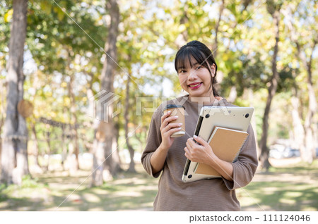 A beautiful young Asian woman stands in a park with a coffee cup, a laptop, and a book in her hands. 111124046