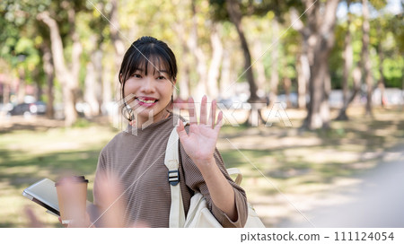 A friendly female student is waving her hand, greeting, or saying goodbye to her friend in a park. A friendly female student is waving her hand, greeting, or saying goodbye to her friend in a park. 111124054