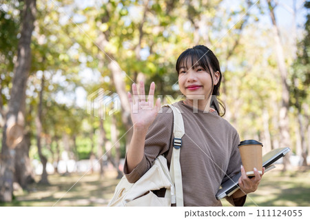 A friendly female student is waving her hand, greeting, or saying goodbye to her friend in a park. A friendly female student is waving her hand, greeting, or saying goodbye to her friend in a park. 111124055