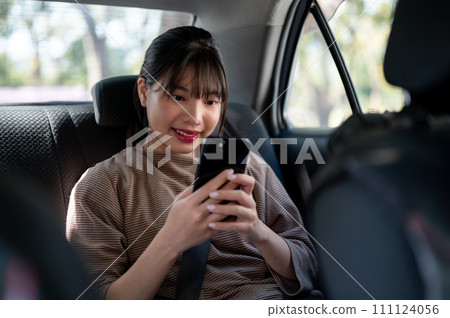 A happy young Asian female passenger is using her smartphone in a taxi, chatting with someone. A happy young Asian female passenger is using her smartphone in a taxi, chatting with someone. 111124056
