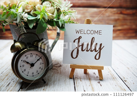 Welcome July text message written on paper card with wooden easel and alarm clock with flower in metal vase decoration 111126497