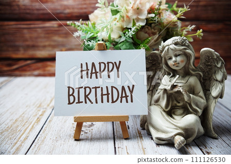 Happy Birthday text message written on paper card with wooden easel and angel statue with flower in metal vase decoration 111126530
