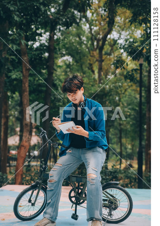 Young handsome bearded man taking a break while travelling the city with his bicycle using his digital tablet looking away thoughtfully Young handsome bearded man taking a break while travelling the city with his bicycle using his digital tablet looking away thoughtfully 111128158