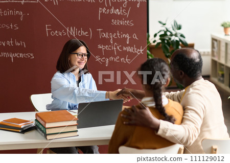 Portrait of young woman as teacher or student advisor shaking hands with parent during meeting in class 111129012