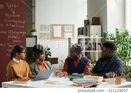 Portrait of senior Black professor talking to group of students sitting at table together in college classroom copy space Portrait of senior Black professor talking to group of students sitting at table together in college classroom copy space 111129057