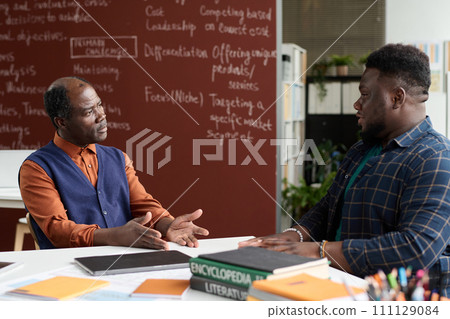 Portrait of Black senior professor listening to male student during one on one consultation in school classroom Portrait of Black senior professor listening to male student during one on one consultation in school classroom 111129084