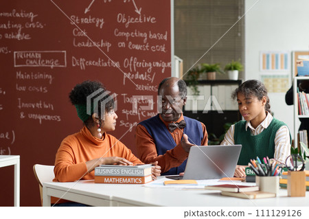 Front view portrait of senior Black teacher talking to students at table in college classroom and pointing to laptop screen copy space 111129126