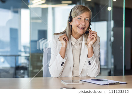 Happy elderly female in light formal clothes in wireless headset setting microphone by table in office space. Blue eyed woman working as customer support representative and providing service. 111133215