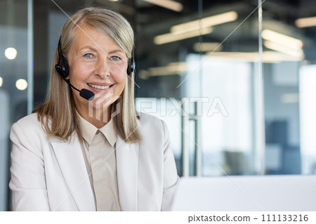 Portrait of mature lady in headset working by computer and smiling at camera on blurred background. Progressive entrepreneur using wireless technology while waiting for connecting to conference. Portrait of mature lady in headset working by computer and smiling at camera on blurred background. Progressive entrepreneur using wireless technology while waiting for connecting to conference. 111133216