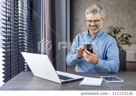 Satisfied middle aged freelancer using mobile phone by desktop with computer next to window with jalousie. Smiling relaxed worker reading pleasant text message during busy day at home workspace. Satisfied middle aged freelancer using mobile phone by desktop with computer next to window with jalousie. Smiling relaxed worker reading pleasant text message during busy day at home workspace. 111133226