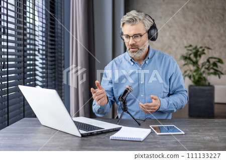 Focused aged male in headphones sitting by working table with laptop and professional microphone indoors. Busy bearded man recording podcast lecture while using modern technology for best acoustic. 111133227
