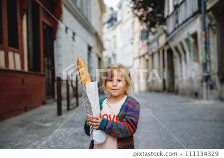 Adorable little preschool girl with fresh French baguette on the street side of the city. Happy small child in Paris, France. 111134329