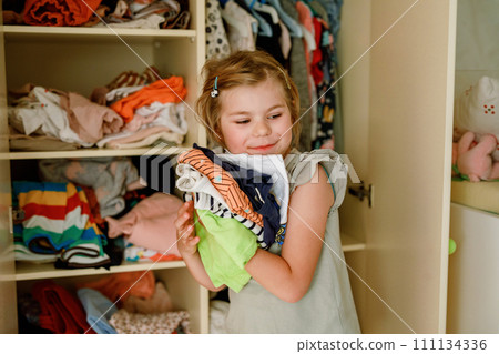 Little school girl standing by wardrobe with clothes. Preschool child putting shirts away and making decision for shirt to wear. Children get dressed in the morning for school. 111134336