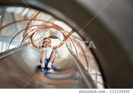 Little preschool girl riding from childrens slides on playground. Happy smiling child having fun in game center for kids. 111134342