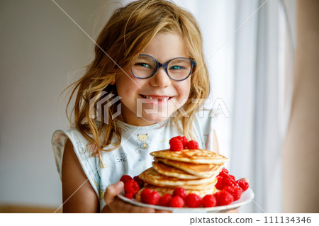 Little happy preschool girl with a large stack of pancakes and raspberries for breakfast. Positive child eating healthy homemade food in the morning. Little happy preschool girl with a large stack of pancakes and raspberries for breakfast. Positive child eating healthy homemade food in the morning. 111134346