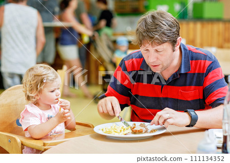 Toddler girl eating healthy vegetables and unhealthy french fries potatoes. Cute happy baby child taking food from parents dish in restaurant. Father eating in fast food restaurant with daughter 111134352
