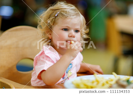 Adorable toddler girl eating healthy vegetables and unhealthy french fries potatoes. Cute happy baby child taking food from dish at daycare or nursery canteen. 111134353