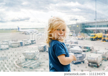 Cute little toddler girl at the airport, traveling. Happy healthy child waiting near window and watching airplanes. Family going on summer vacations by plane. 111134375