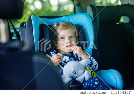 Adorable baby girl with blue eyes sitting in car safety seat. Toddler child going on family vacations and jorney. Smiling happy child during traffic jam, eating bisquit 111134397