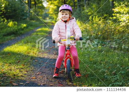 Child riding balance bike. Kids on bicycle in sunny forest. Little girl enjoying to ride glider bike on warm day. Preschooler learning to balance on run bicycle in safe helmet. Sport activity 111134436