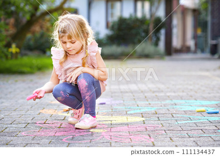 Little preschool girl painting with colorful chalks flowers on ground on backyard. Positive happy toddler child drawing and creating pictures on asphalt. Creative outdoors children activity in summer. Little preschool girl painting with colorful chalks flowers on ground on backyard. Positive happy toddler child drawing and creating pictures on asphalt. Creative outdoors children activity in summer. 111134437
