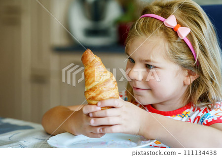 Smiling child at breakfast. Food and happy kids. The girl is eating a croissant. Cute preschool girl having healthy meal. 111134438