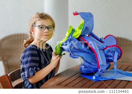 Happy smiling girl preparing for school her backpack. First day of school. Back to school concept. Little child collecting different supplies like pens and books in a bag or satchel. 111134491