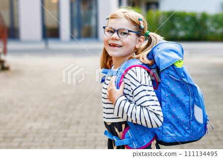 Cute little girl on her first day going to school. Healthy beautiful child walking to nursery preschool and kindergarten. Happy child with eyeglasses with backpack on the city street, outdoors. Cute little girl on her first day going to school. Healthy beautiful child walking to nursery preschool and kindergarten. Happy child with eyeglasses with backpack on the city street, outdoors. 111134495