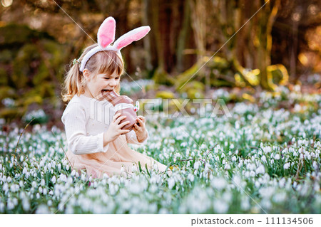 Happy little girl with Easter bunny ears eating chocolate figure in spring forest on sunny day, outdoors. Cute child with lots of snowdrop flowers. Springtime, christian holiday concept. Happy little girl with Easter bunny ears eating chocolate figure in spring forest on sunny day, outdoors. Cute child with lots of snowdrop flowers. Springtime, christian holiday concept. 111134506