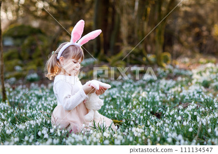 Adorable little girl with Easter bunny ears holding soft plush toy in spring forest on sunny day, outdoors. Cute happy child with lots of snowdrop flowers. Springtime, christian holiday concept. 111134547