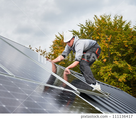 Builder mounting photovoltaic solar panels on roof of house. Man engineer in helmet installing solar modules system outdoors. Concept of alternative and renewable energy. 111136417