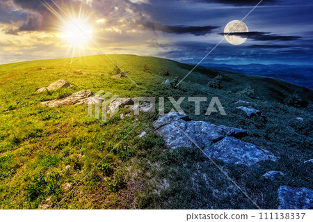 mountain landscape with stones among the grass on top of the hillside beneath a sky with sun and moon at twilight. day and night time change concept. mysterious countryside scenery in morning light 111138337